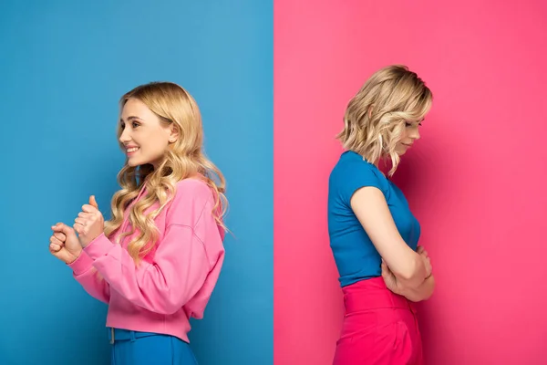 Side view of sad blonde girl near sister showing yes gesture on pink and blue background — Stock Photo