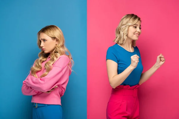 Sad blonde girl looking away near happy sister showing yeah gesture on pink and blue background — Stock Photo