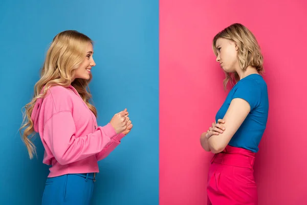 Vista lateral de hermanas rubias tristes y felices mirándose sobre fondo rosa y azul - foto de stock