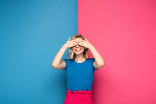 Lächelndes blondes Mädchen, das Augen auf rosa und blauem Hintergrund bedeckt — Stockfoto