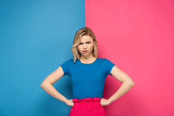 Wütendes blondes Mädchen mit den Händen auf den Hüften, das in die Kamera auf rosa und blauem Hintergrund blickt — Stockfoto