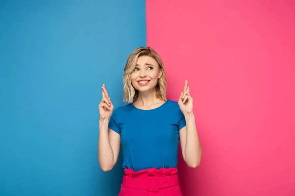 Fille blonde souriante avec les doigts croisés sur fond rose et bleu — Photo de stock