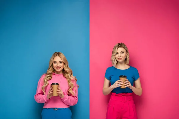 Smiling blonde sisters holding paper cups on pink and blue background — Stock Photo