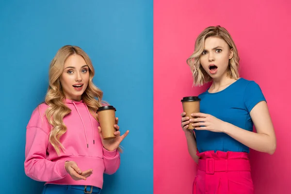 Surprises femmes blondes avec des tasses jetables regardant la caméra sur fond rose et bleu — Photo de stock