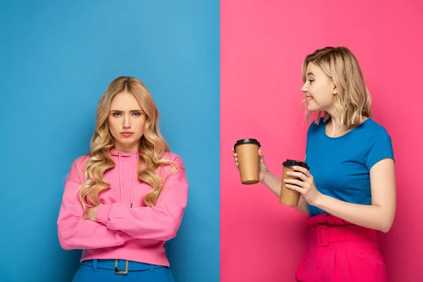 Smiling blonde girl with paper cups looking at offended sister on pink and blue background — Stock Photo