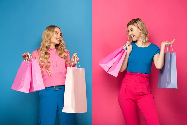 Chicas rubias sonrientes con bolsas de compras mirándose el uno al otro sobre fondo rosa y azul - foto de stock