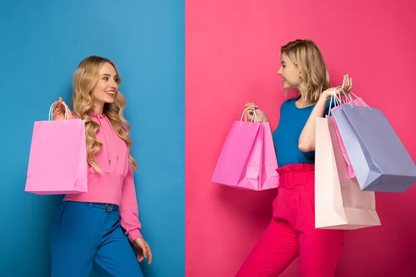 Attractive blonde sisters holding shopping bags and smiling at each other on pink and blue background — Stock Photo