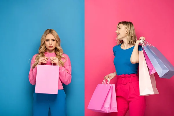 Menina sorridente com sacos de compras olhando para a irmã ofendida no fundo rosa e azul — Fotografia de Stock