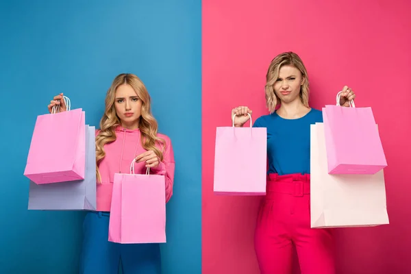Beleidigte blonde Mädchen halten Einkaufstüten in der Hand und lächeln in die Kamera auf rosa und blauem Hintergrund — Stockfoto