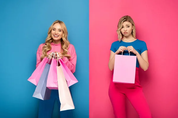 Усміхнена блондинка з сумками дивиться на камеру поблизу ображеної сестри на рожевому і синьому фоні — стокове фото
