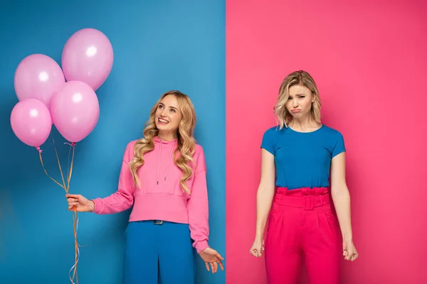 Smiling blonde girl holding balloons near sad sister on blue and pink background — Stock Photo