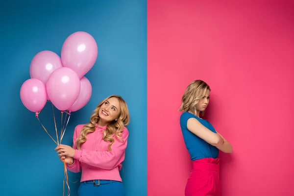 Lächelndes blondes Mädchen mit Luftballons neben trauriger Schwester mit verschränkten Armen auf blauem und rosa Hintergrund — Stockfoto