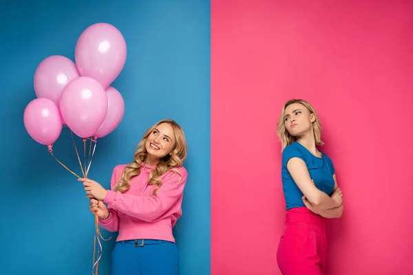 Offended blonde girl looking at cheerful sister with balloons on blue and pink background — Stock Photo