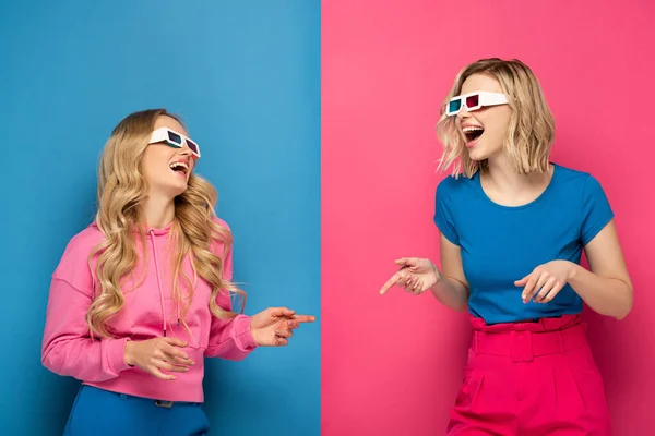 Lachende blonde Schwestern in 3D-Brille, die mit den Fingern auf blauem und rosa Hintergrund zeigen — Stockfoto