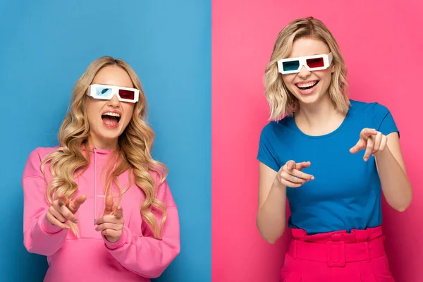 Irmãs loiras positivas em óculos 3d apontando com os dedos para a câmera em fundo azul e rosa — Fotografia de Stock