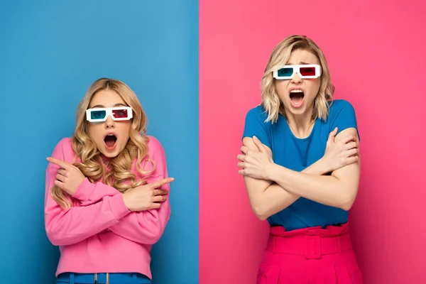 Scared blonde sisters in 3d glasses looking at camera on blue and pink background — Stock Photo