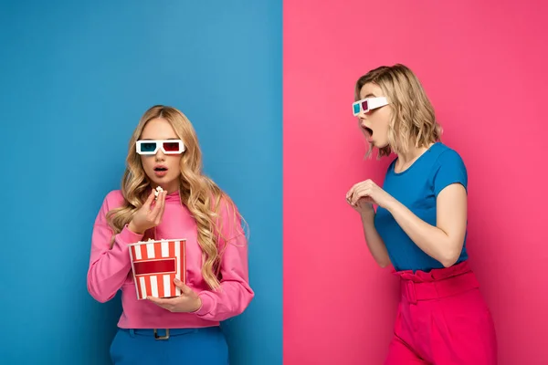 Sorprendido chica rubia en gafas 3d mirando hermana comiendo palomitas de maíz sobre fondo azul y rosa - foto de stock