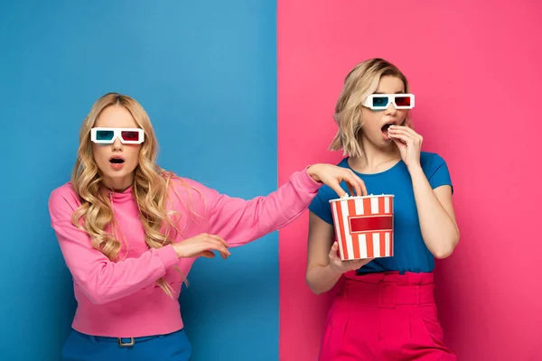 Excited blonde woman in 3d glasses taking popcorn near sister on blue and pink background — Stock Photo