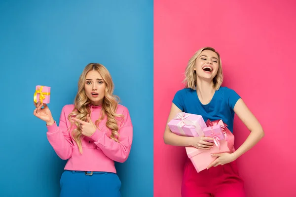 Заплутана блондинка вказує на подарункову коробку біля позитивної сестри, що тримає подарунки на синьо-рожевому фоні — стокове фото
