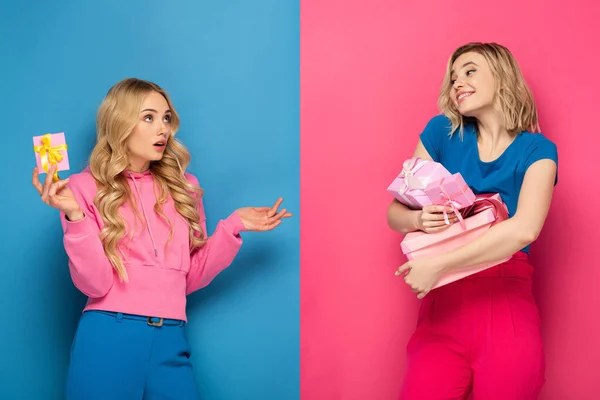 Confused blonde girl holding present near smiling sister with gift boxes on blue and pink background — Stock Photo