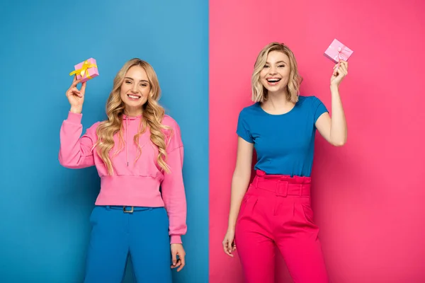 Filles blondes souriantes tenant des cadeaux et regardant la caméra sur fond bleu et rose — Photo de stock