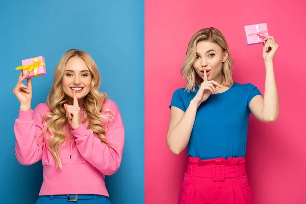 Positive blonde sisters holding gift boxes and showing silence sign on blue and pink background — Stock Photo