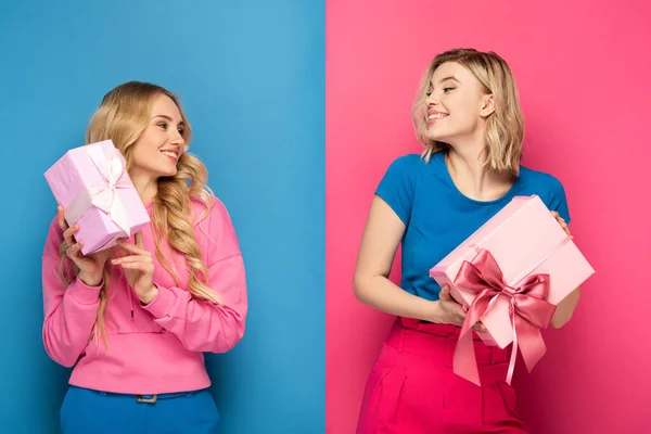 Lächelnde blonde Mädchen schauen einander an, während sie Geschenkboxen auf blauem und rosa Hintergrund halten — Stockfoto