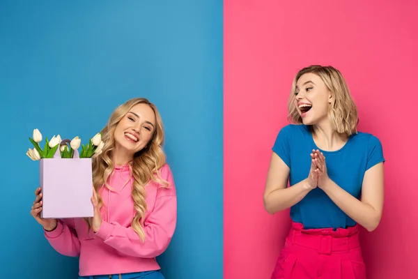 Joyeux soeur blonde avec bouquet dans un sac à provisions sur fond rose et bleu — Photo de stock