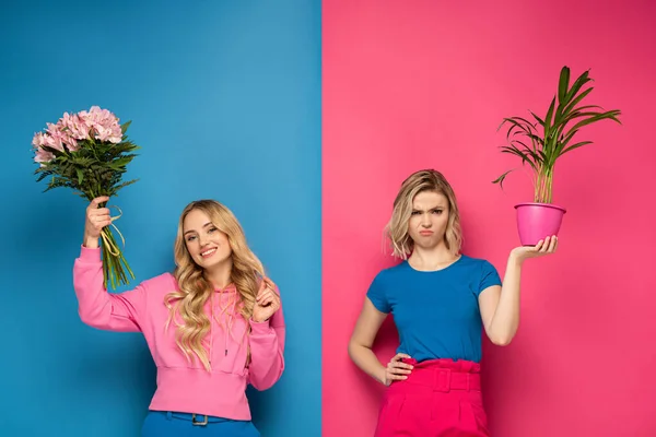 Sonriente chica sosteniendo ramo cerca ofendida hermana con planta sobre fondo rosa y azul - foto de stock