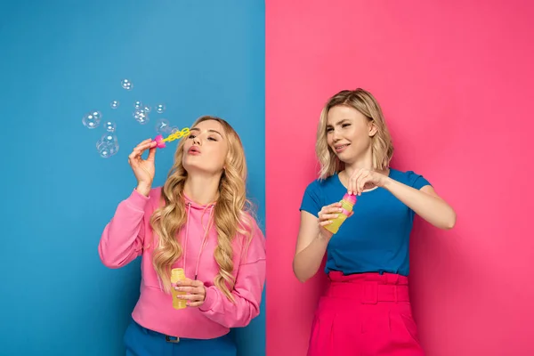 Beautiful blonde girl blowing soap bubbles near skeptical sister on pink and blue background — Stock Photo