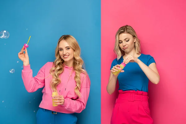 Smiling blonde girl holding soap bubbles near sister on pink and blue background — Stock Photo