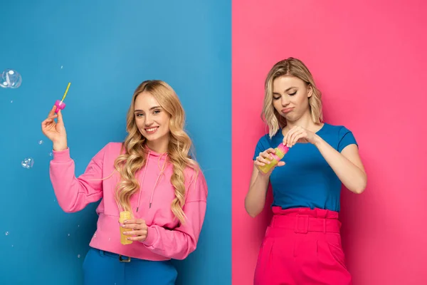 Sonriente chica rubia sosteniendo burbujas de jabón cerca de la hermana sobre fondo rosa y azul - foto de stock