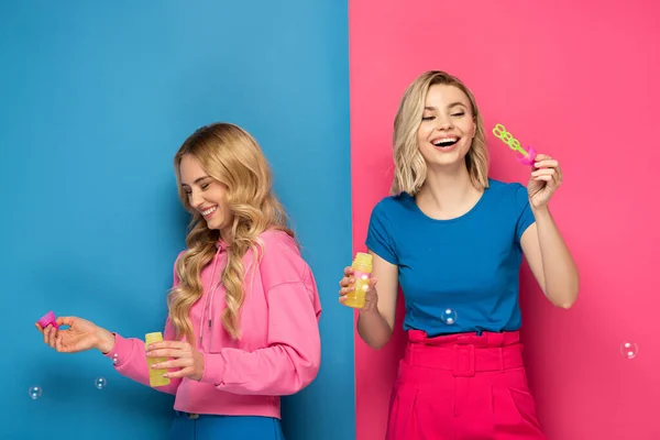 Happy blonde women blowing soap bubbles on pink and blue background — Stock Photo