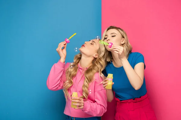 Beautiful blonde sisters blowing soap bubbles on pink and blue background — Stock Photo