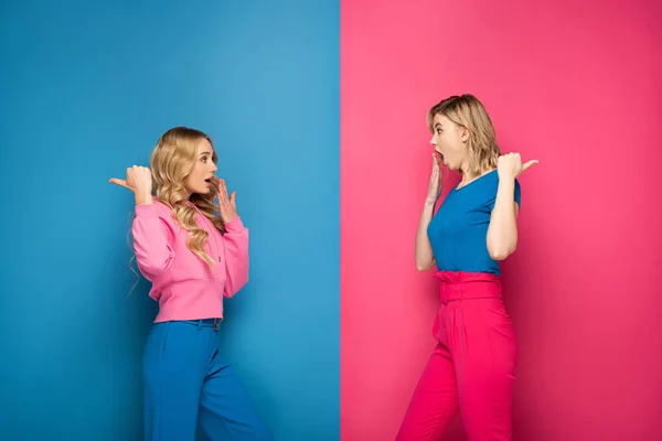 Chicas rubias impactadas señalando con los dedos sobre fondo rosa y azul - foto de stock