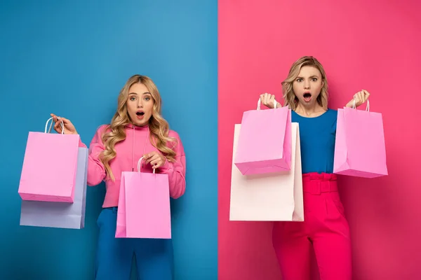 Surprised blonde sisters with shopping bags looking at camera on pink and blue background — Stock Photo