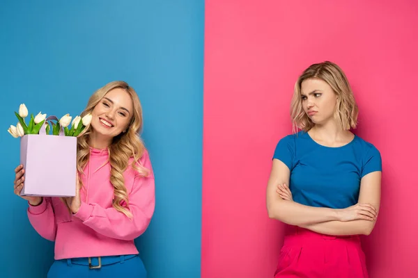 Smiling girl holding bouquet near envy blonde sister on blue and pink background — Stock Photo