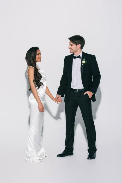 Vista completa de feliz, elegante interracial recién casados tomados de la mano y mirándose el uno al otro sobre fondo blanco - foto de stock