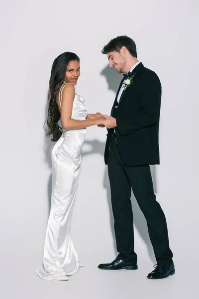 Vue pleine longueur de heureuse, belle mariée afro-américaine regardant la caméra tout en tenant la main avec marié élégant sur fond blanc — Photo de stock