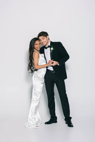 Vista completa de feliz, elegante interracial recién casados cogidos de la mano y sonriendo a la cámara sobre fondo blanco - foto de stock