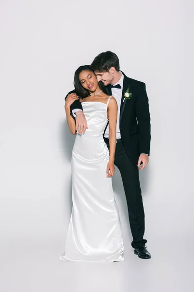 Full length view of happy, elegant interracial newlyweds smiling while posing on white background — Stock Photo