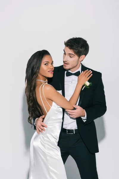 Elegante, atractiva novia afroamericana mirando a la cámara mientras toca novio guapo sobre fondo blanco - foto de stock