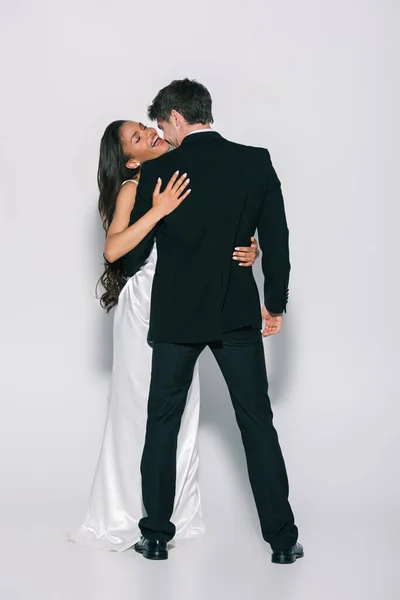 Vue pleine longueur de heureuse mariée afro-américaine embrassant marié élégant sur fond blanc — Photo de stock