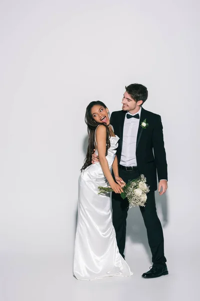 Vue pleine longueur de marié élégant étreignant heureuse mariée afro-américaine tenant bouquet de mariage sur fond blanc — Photo de stock