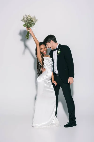 Visão de comprimento total da noiva americana africana feliz segurando buquê de casamento na mão levantada perto do noivo sorridente no fundo branco — Fotografia de Stock