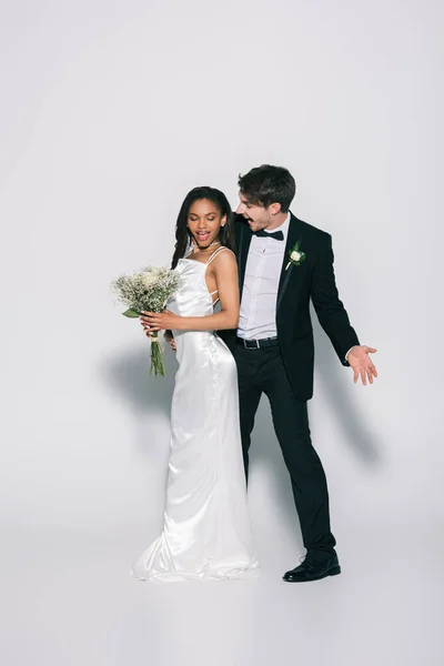 Vue pleine longueur de marié excité regardant élégante mariée afro-américaine tenant bouquet de mariage sur fond blanc — Photo de stock