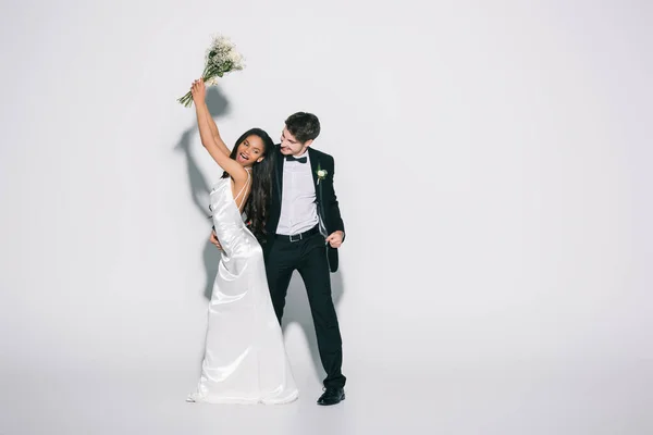 Full length view of happy african american bride holding wedding bouquet in raised hands near elegant bridegroom on white background — Stock Photo