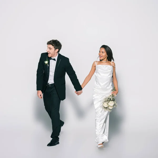 Vista completa de sonrientes y elegantes recién casados interracial tomados de la mano mientras caminan y miran hacia otro lado sobre fondo blanco - foto de stock