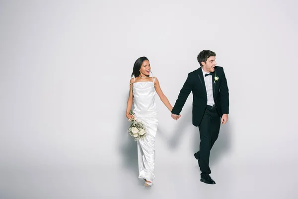 Full length view of happy, elegant interracial newlyweds holding hands while walking and looking away on white background — Stock Photo