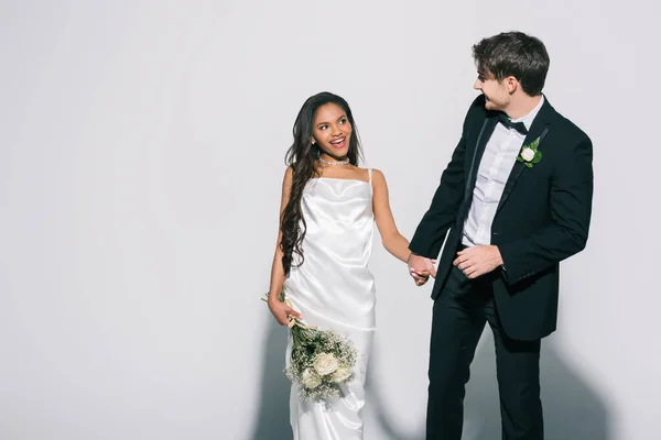 Noivo de noiva elegante segurando a mão da noiva americana africana feliz com buquê de casamento no fundo branco — Fotografia de Stock